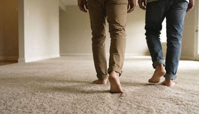 Couple walking barefoot across plush beige carpet in empty apartment, rolling pant cuffs