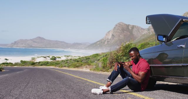 Man sitting on road next to broken down car along scenic coastal route with mountainous backdrop. Ideal for use in content about travel issues, car maintenance, or emotional reflection during journeys.