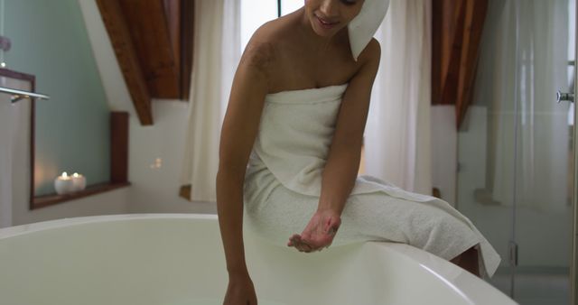 Woman sitting by a bathtub in a modern, cozy bathroom with wooden ceiling and natural light. She is dressed in a bathrobe and a towel on her head, checking water temperature. Background includes lit candles, enhancing the relaxing atmosphere. Perfect for use in content related to wellness, self-care, relaxation, spa treatments, and interior design promotions.