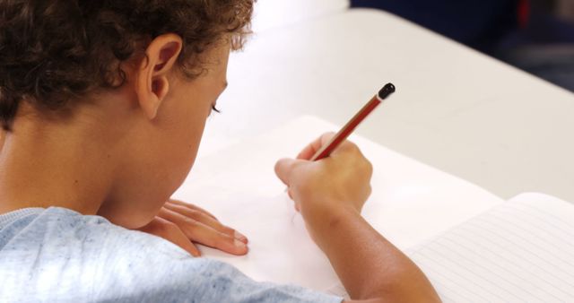 A child with curly hair is writing in a notebook, focused on his homework in a classroom. Useful for educational content, school-related materials, advertisements for education tools or tutoring services, and parenting advice articles.