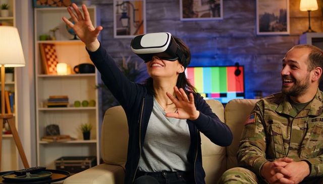 Woman experiencing virtual reality while soldier partner watches laughing in cozy living room