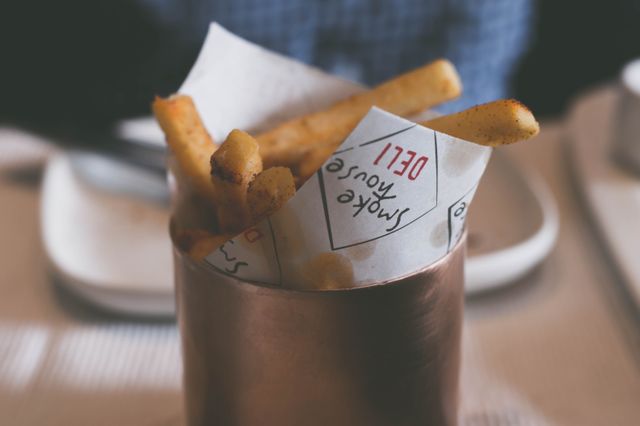 Crispy French fries served in a rustic metal cup with deli branding at a smoke house restaurant. Ideal for use in culinary blogs, restaurant promotions, food magazines, and social media posts related to dining, fast food, and savory snacks.
