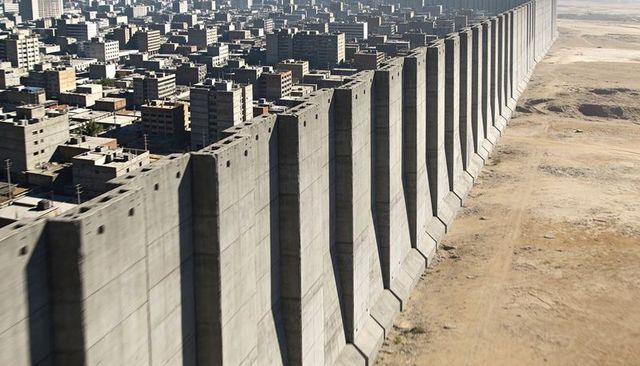 Massive concrete wall dividing dense city from desert plain with rhythmic buttresses