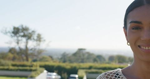 Smiling woman wearing crochet top and hoop earring in sunlit garden portrait, driveway view