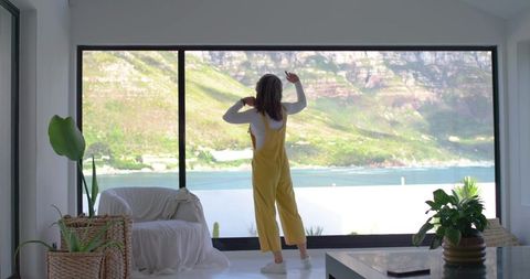 Woman in yellow overalls standing by panoramic coastal window in minimalist bright interior