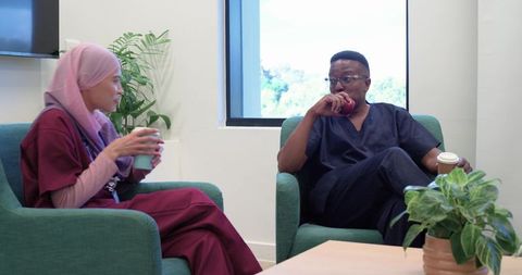 African american nurse and colleague in hijab taking break in hospital lounge eating apple