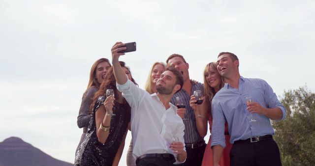 Group of friends smiling and posing for selfie during outdoor celebration. People dressed casually with wine glasses in hand enjoying social gathering. Use for content related to social events, friendships, parties, celebrations, outdoor activities, and lifestyle imagery.