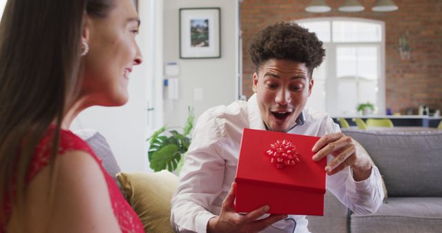 Man and woman wearing casual clothes with man holding a red gift box and expressing surprise and joy, sitting on a couch in a bright, modern living room. Suitable for themes like celebrations, gifting, emotions, romantic occasions, and special moments within relationships.