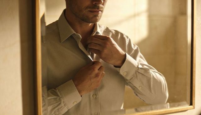 Man buttoning dress shirt in warm mirror light preparing for work and evening occasion