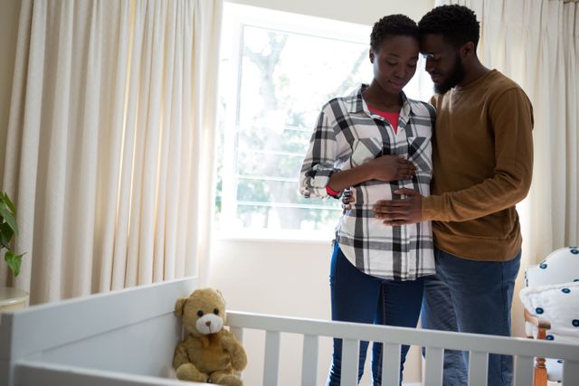 Expecting parents embracing in a nursery, with the man touching the pregnant woman's stomach. The scene includes a baby crib and a teddy bear, creating a warm and loving atmosphere. This image is perfect for use in parenting blogs, maternity articles, family planning resources, and advertisements for baby products.