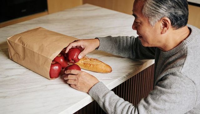 Asian man unpacking groceries on marble counter reaching for red apples and baguette in cozy kitchen