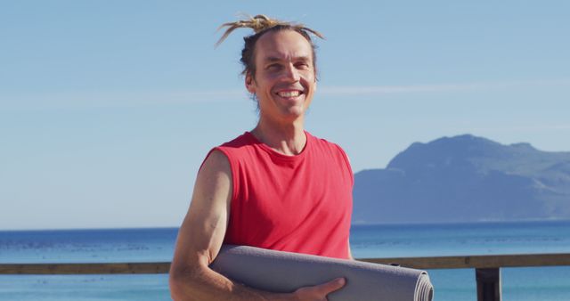 Man holding a rolled-up yoga mat while smiling joyfully on a beautiful beach with mountains in the background. Ideal for advertisements promoting outdoor activities, yoga retreats, fitness, healthy lifestyles, and travel experiences. Perfect for use in websites, blogs, and magazines focused on wellness and outdoor exploration.