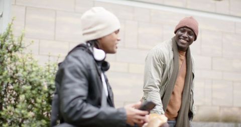 Diverse young men chatting and laughing on urban bench with smartphone and sandwich