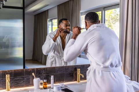 Mature man brushing teeth at bathroom vanity in white bathrobe with mirror reflection