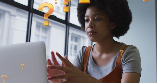 African American woman sitting at desk in office, looking frustrated and confused while using laptop, surrounded by question marks. Useful for illustrating concepts of work stress, problem-solving, technology challenges, and confusion in a professional environment.