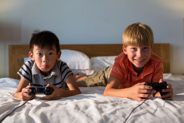 Two boys are lying on a bed, holding video game controllers and playing a video game. They are smiling and appear to be enjoying their time together. This image can be used for themes related to childhood, sibling bonding, indoor activities, and technology in everyday life.