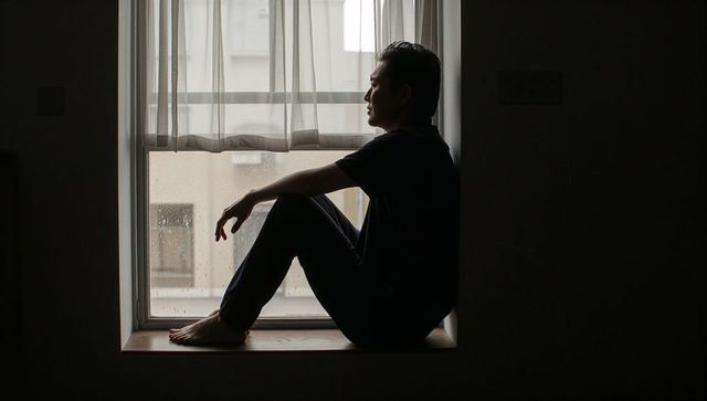Sitting barefoot man gazing through rainy window silhouette on windowsill, moody interior