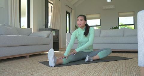 Mid-adult asian woman performing seated hamstring stretch at home in mint activewear on yoga mat
