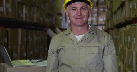 Smiling warehouse supervisor wearing yellow hard hat standing in pallet racking aisle with laptop