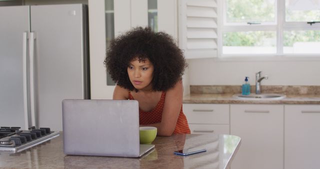 Woman using a laptop on kitchen countertop, demonstrating multitasking in a home environment. Ideal for use in articles, blog posts, or adverts about remote work, home offices, technology use, or modern kitchens.