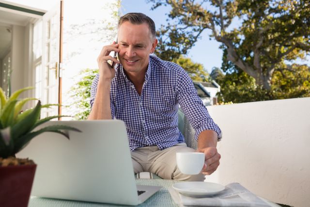 Mature businessman enjoying coffee while talking on phone and working on laptop outdoors. Ideal for illustrating remote work, business communication, professional lifestyle, and multitasking in a relaxed environment.