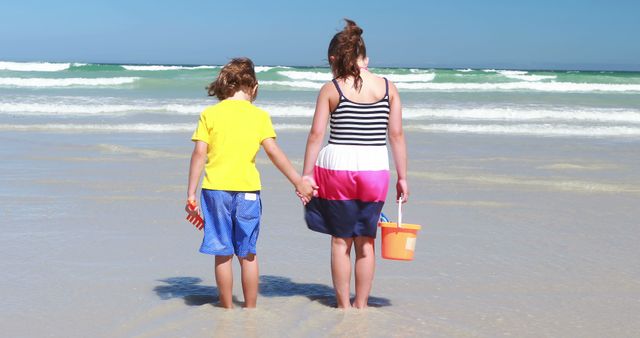 A young boy and girl hold hands while standing on a sandy beach, facing the ocean, with copy space. Their casual beach attire and the presence of a bucket suggest a playful day spent by the sea.