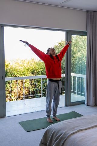 Asian man stretching on yoga mat in bedroom near balcony morning routine red hoodie