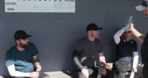 Baseball teammates sitting in dugout holding water bottles and glove under sunlight