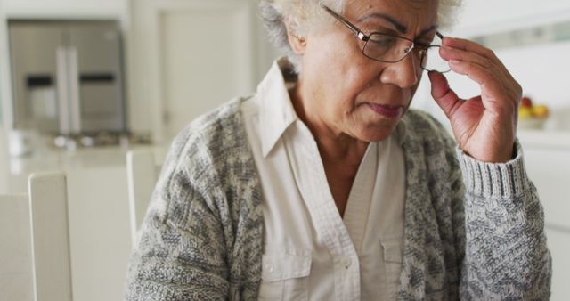Elderly woman with grey hair, wearing glasses and a sweater, appears thoughtful at home. Can be used in contexts related to aging, senior living, elder care, and the mental health of the elderly.