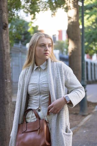 Young woman standing on tree-lined street wearing cardigan, carrying tan leather bag