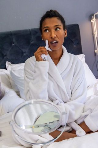 African american woman using facial skincare device in white bathrobe on bed
