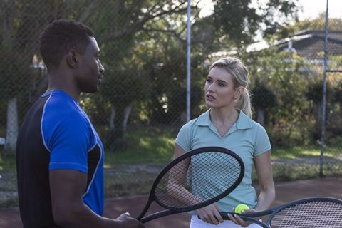 Black man and white woman practicing tennis on outdoor court, talking and holding rackets
