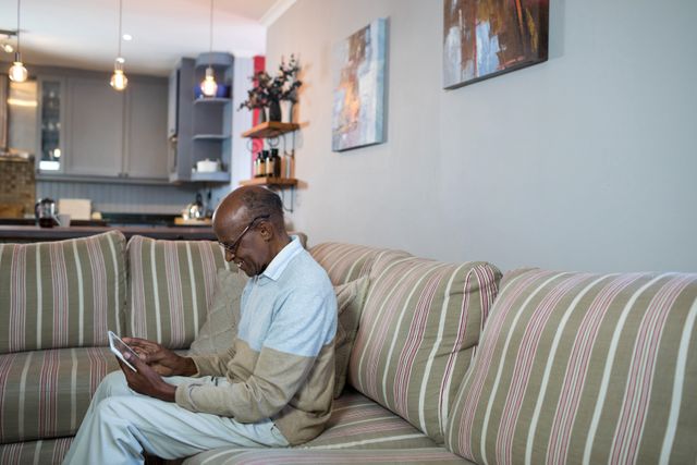 Senior man sitting on a striped sofa in a modern living room, using a tablet. Ideal for topics related to technology use among seniors, retirement lifestyle, home comfort, and digital literacy.
