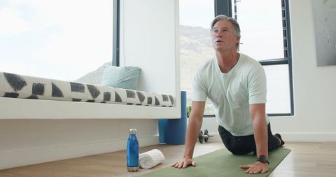 Mature man practicing morning yoga on mat by window with water bottle and dumbbell