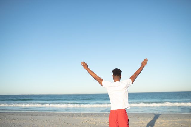 Man standing on beach with arms raised, facing ocean. Ideal for themes of freedom, relaxation, vacation, and personal achievement. Perfect for travel brochures, wellness blogs, and motivational content.