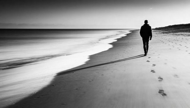 Solitary man walking along empty monochrome beach leaving footprints and long shadow