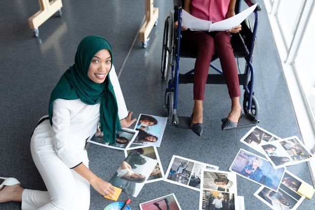 Two female graphic designers, one wearing a hijab and the other in a wheelchair, are reviewing photographs in a modern office. This image can be used to represent diversity, inclusion, and collaboration in a creative work environment. Ideal for articles or advertisements focusing on inclusive workplaces, creative industries, and teamwork.