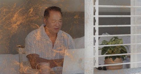 Mature chinese man working on laptop in bedroom workspace with mug and indoor plant