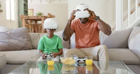 Father and son playing virtual reality games on modern sofa with headsets and controllers