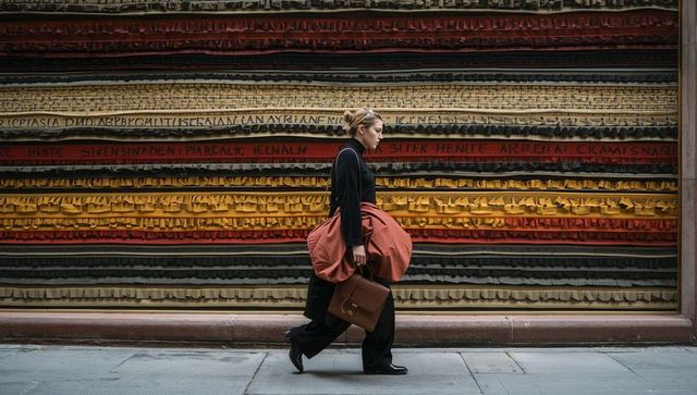 Striding woman passing banded shutter wearing tied orange coat carrying leather bag