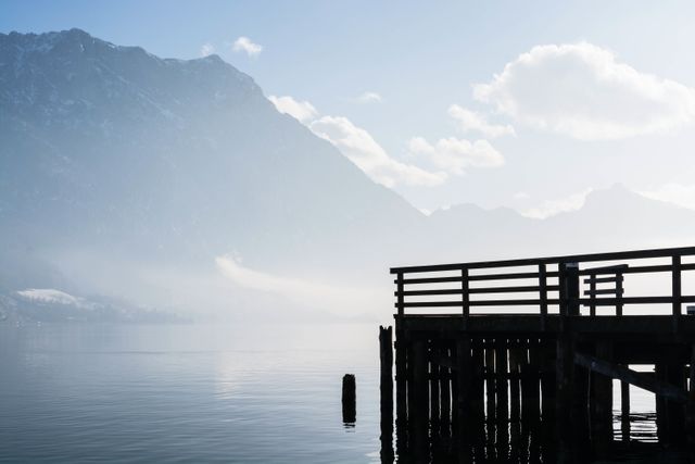 This image captures a tranquil moment of a foggy lake with a wooden pier, set against the serene backdrop of picturesque mountains. Ideal for uses related to travel, outdoor activities, relaxation, meditation visuals, nature blogs, and scenic photography collections.