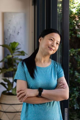 Mature asian woman standing leaning on sliding glass door, arms crossed, wearing smartwatch