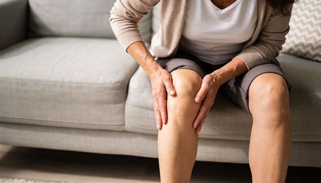 Senior woman holding right knee while sitting on gray sofa, joint pain and mobility focus