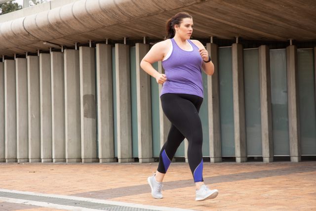 Curvy Caucasian woman with long dark hair wearing sports clothes exercising in a city, running past modern architecture in an urban pedestrian area