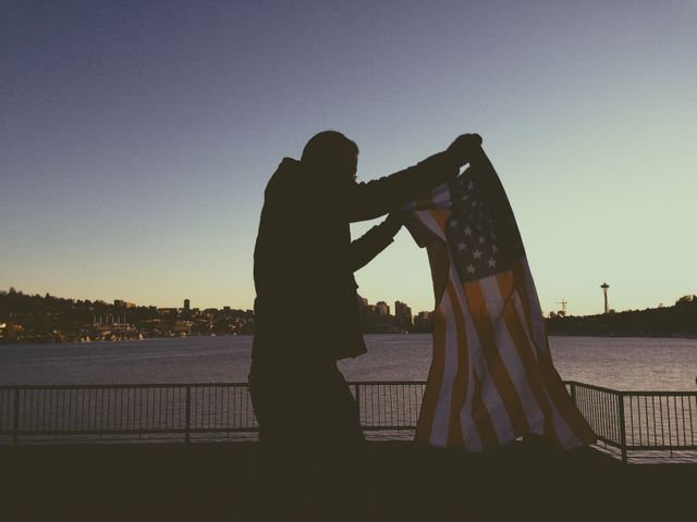 Depicting a silhouette of a person holding an American flag by a waterfront during sunset. Can be used for themes of patriotism, freedom, national pride, and reflection. Suitable for social media posts, blog illustrations, and editorial content related to national holidays or American culture.