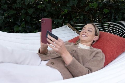 Relaxing woman making mobile payment while reclining on white hammock in backyard garden
