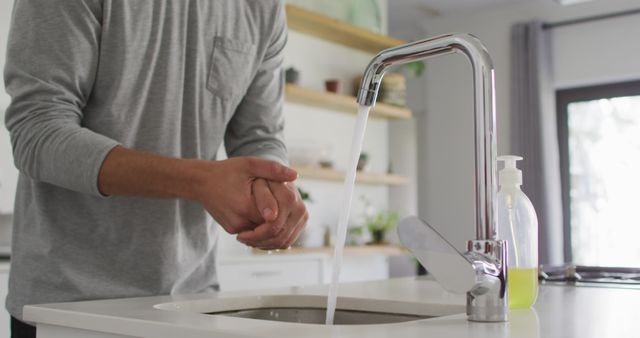 Closeup of a person washing hands under running water in a modern kitchen. Soap dispenser and tap are visible. Perfect for use in hygiene promotions, health and wellness articles, and modern home editorial content.