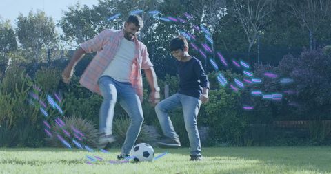Father and son playing soccer on sunlit lawn with colorful motion light streaks