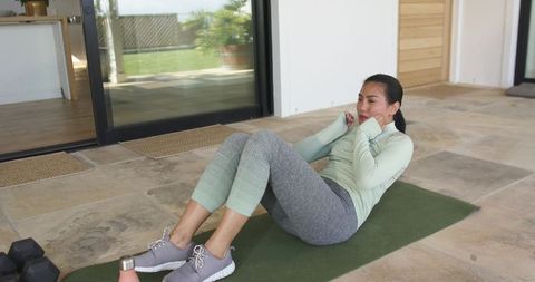 Asian woman performing core workout on green mat on tiled patio with dumbbells
