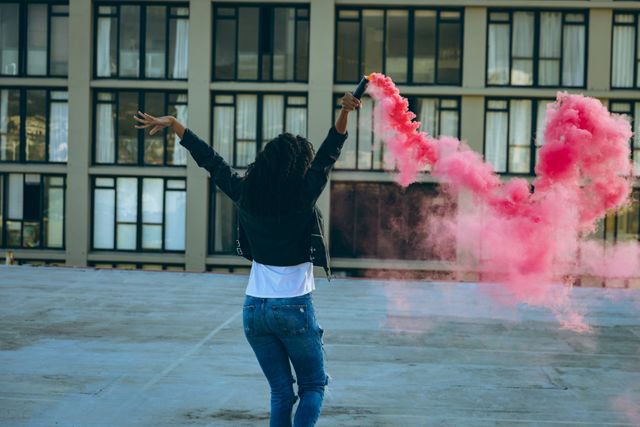 Rear view of a hip young biracial woman wearing leather jacket and jeans, holding a smoke grenade with pink smoke on an urban rooftop with building in the background